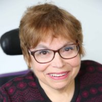 A headshot of Judy Heumann, a cis-gender white woman who is a wheelchair user with short brown hair. She is wearing brown glasses, a maroon and black embroidered cardigan with the top buttoned, and a matching maroon shirt. She is smiling kindly.