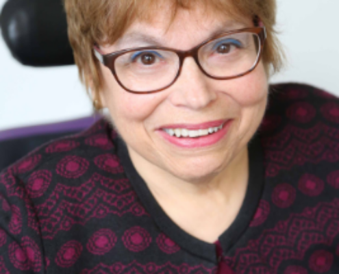 A headshot of Judy Heumann, a cis-gender white woman who is a wheelchair user with short brown hair. She is wearing brown glasses, a maroon and black embroidered cardigan with the top buttoned, and a matching maroon shirt. She is smiling kindly.