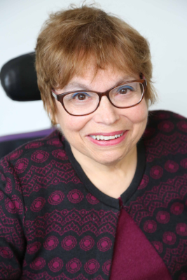 A headshot of Judy Heumann, a cis-gender white woman who is a wheelchair user with short brown hair. She is wearing brown glasses, a maroon and black embroidered cardigan with the top buttoned, and a matching maroon shirt. She is smiling kindly.