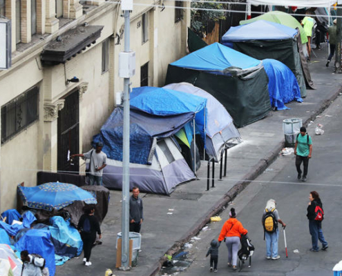 Blue tents set up on the sidewalk of a homeless encampment as some pedestrians walk by.