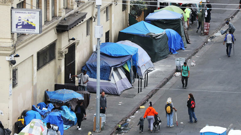 Blue tents set up on the sidewalk of a homeless encampment as some pedestrians walk by.