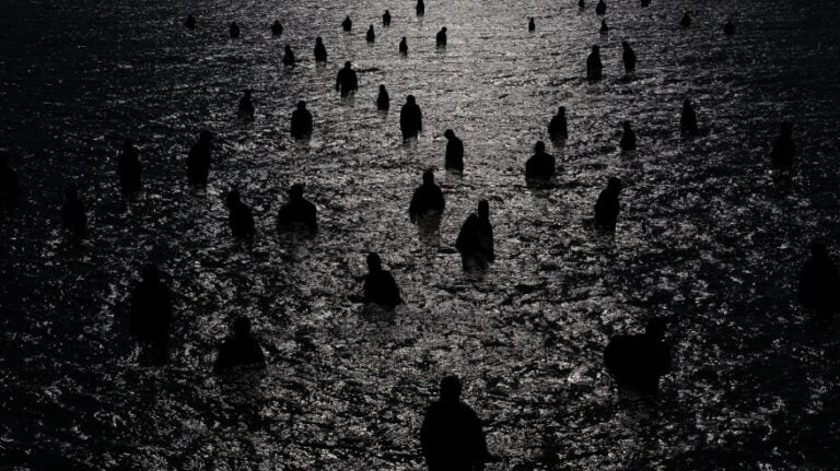 Many dark figures wade in the moonlit water in a black-and-white photo.