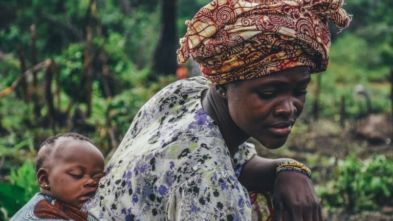 Woman bends down carrying a sleeping baby on her back