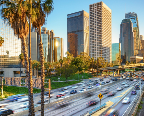 Blurred cars drive by on the freeway with palm trees in the foreground and tall buildings in the background on a sunny Los Angeles day