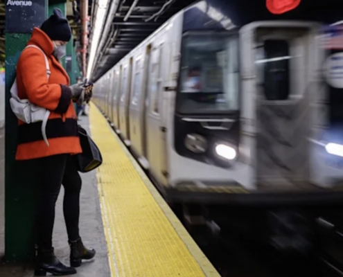 Woman wearing a beanie and a large orange jacket stares at her phone while the subway passes by in the underground station