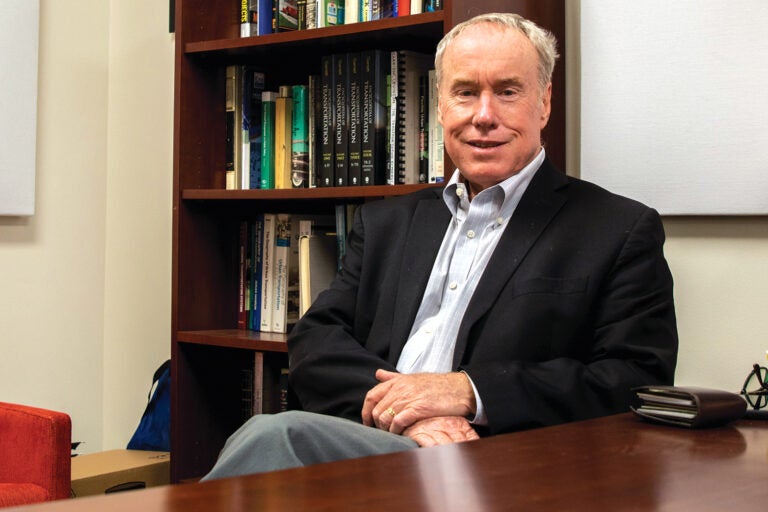 man sits with hands folded on lap and bookshelf in background