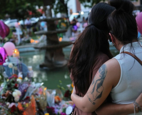 two women embracing in front of memorial of flowers and balloons