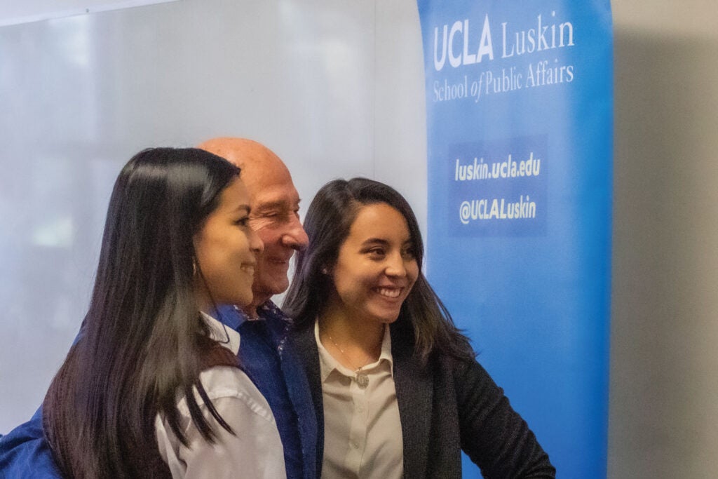 older man poses with two college students at UCLA event|people seated in foreground listen to speaker at podium while a screen shows an image of Martin Wachs|||