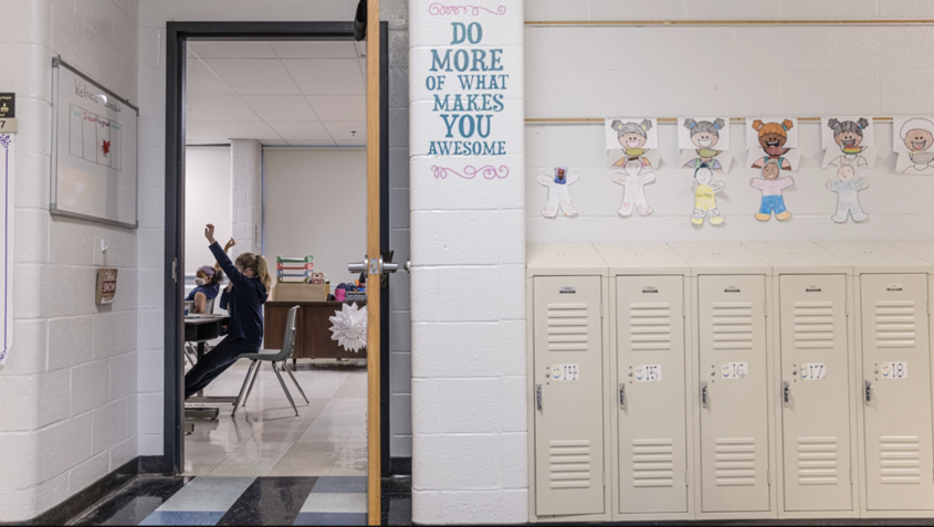Student sitting at a desk in a classroom raises her hand, and a sign posted in the hallway says "Do more of what makes you awesome" next to lockers.