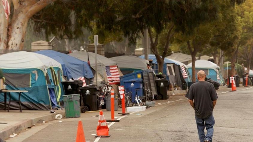 A man walks along the road next to a cluster of tents at a homeless encampment.