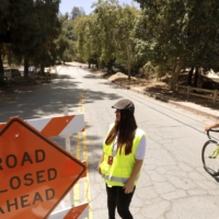 bicyclist on wooded road next to road closure sign and woman in yellow safety vest