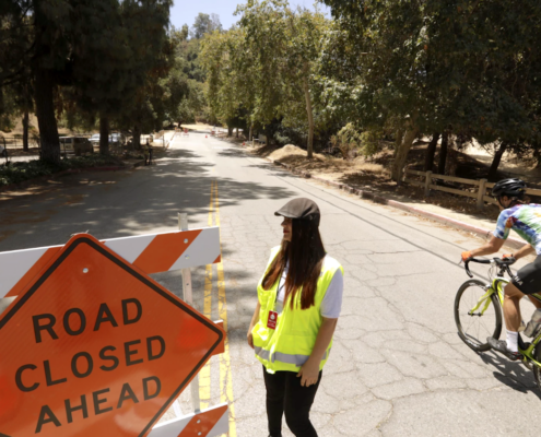 bicyclist on wooded road next to road closure sign and woman in yellow safety vest