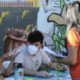 A woman in an orange shirt talks to a man and a woman wearing masks and sitting at a booth in front of a mural at the Healthy Watts Harvest community engagement event in November 2021