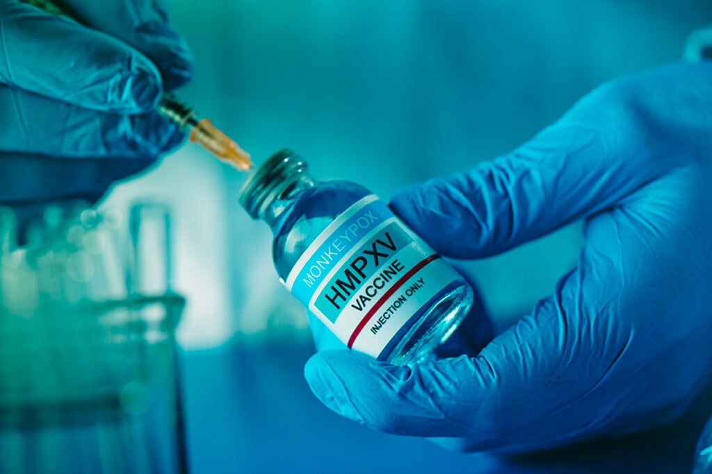 A healthcare or laboratory worker man, wearing blue surgical gloves, fills a syringe with a simulated monkeypox vaccine from a vial|man smiles as he stands beneath sign that designates office location for research hub