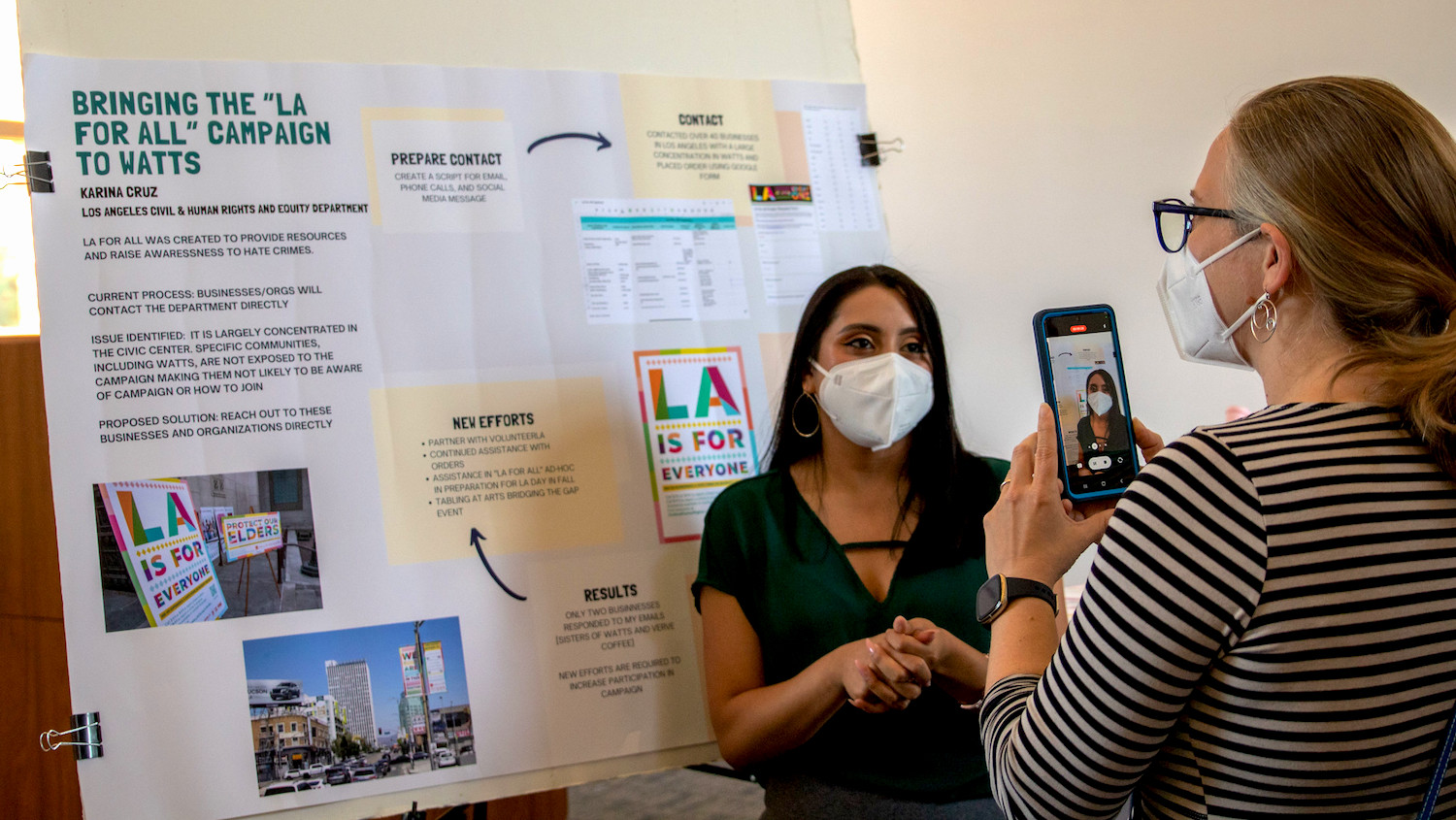 woman in striped shirt takes cellphone video of masked student in front of research poster