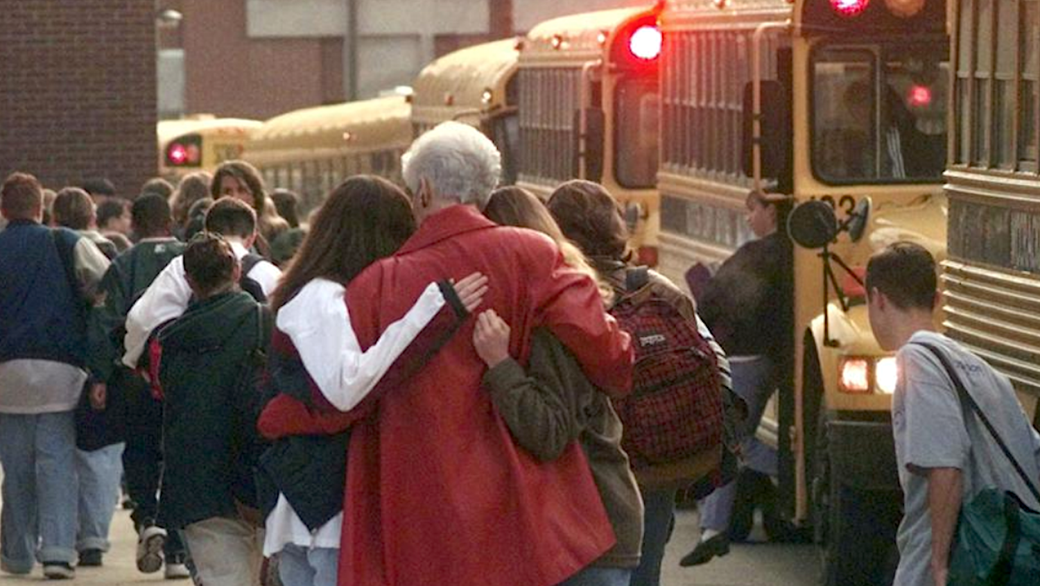 school crowd including woman in red coat hugging two girls in front of school bus