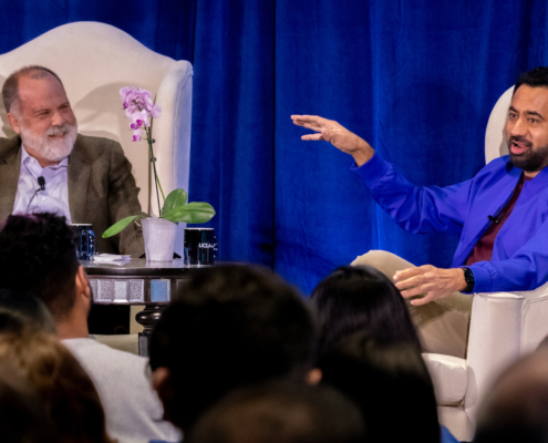 Two men in white chairs on stage in front of blue curtain