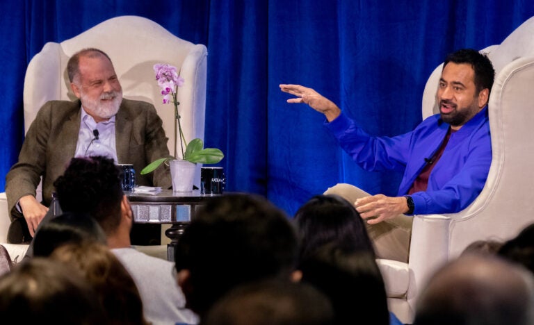 Two men in white chairs on stage in front of blue curtain