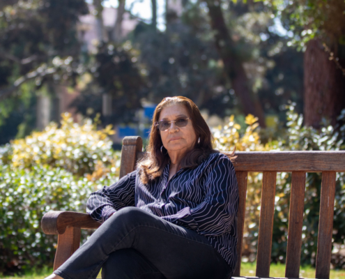 woman in blue top and jeans is surrounded by foliage as she sits on a bench