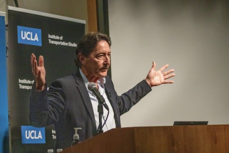 man at podium gestures with arms spread wide|man wearing glasses speaks at podium|woman with gray hair gestures|two men talk while seated in front of a projection screen