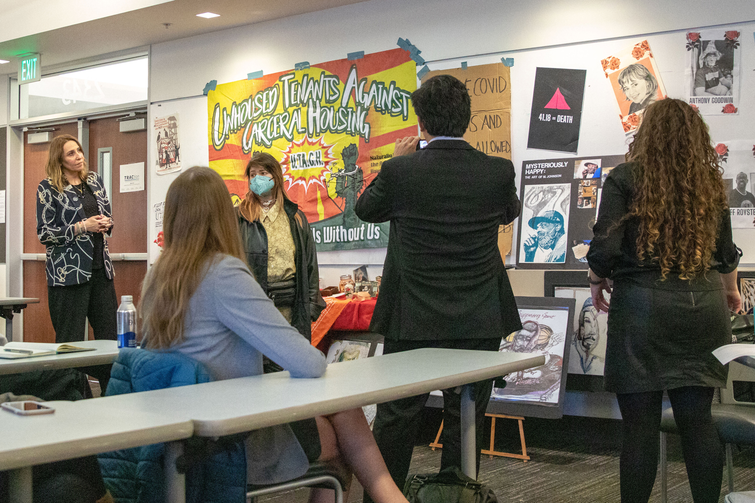 people stand in front of a wall covered with posters, photos and artifacts relating to homelessness