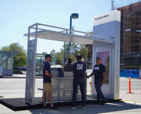 Three people stand underneath a bus shelter on a sunny day.