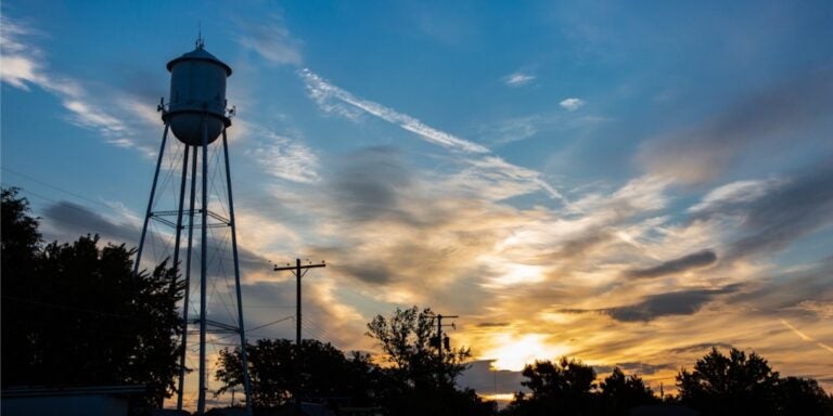 water tower rises over trees with sun just over the horizon in the distance