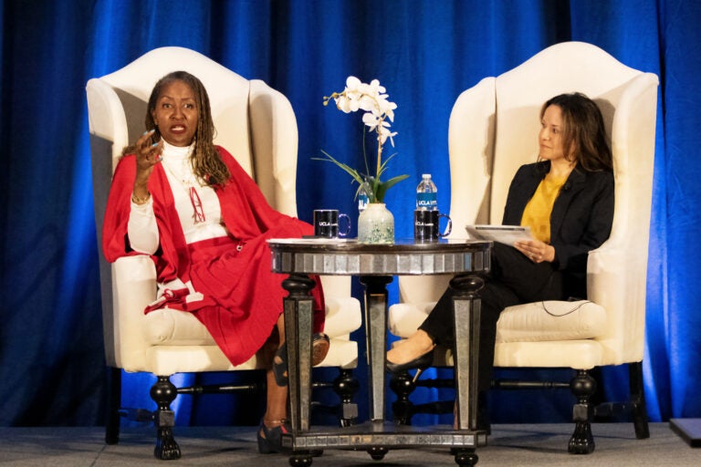 Two women are seated on stage, where a woman in red speaks and the other listens|woman in red shawl at podium gestures forward as she speaks|people seated at tables in a conference room watch a speaker at the front of the room speak at a podium|Two women are seated on stage, where a woman in red speaks and the other listens|woman in purple dress gestures at podium|Black woman leans forward as a woman holding a child speaks with her