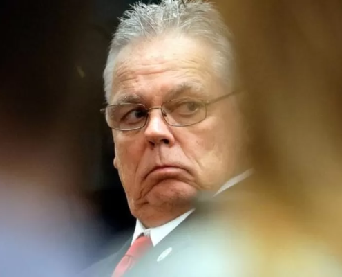 man in glasses and suit looks over his shoulder with a serious expression