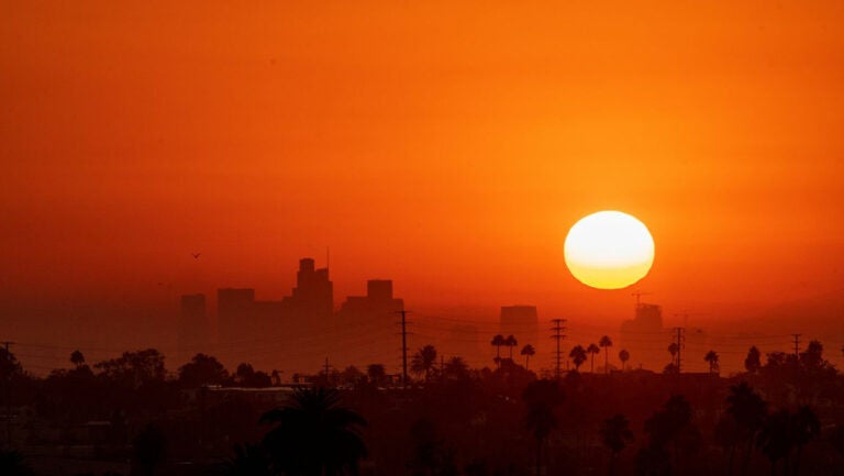 sun hangs low over city with sky looking orange Photo by johnemac72/iStock|woman in striped top speaks at podium while map is projected on a screen behind her|woman in dark shirt without sleeves speaks at a podium|man in blue shirt and glasses gestures at a podium while a woman listens int he background