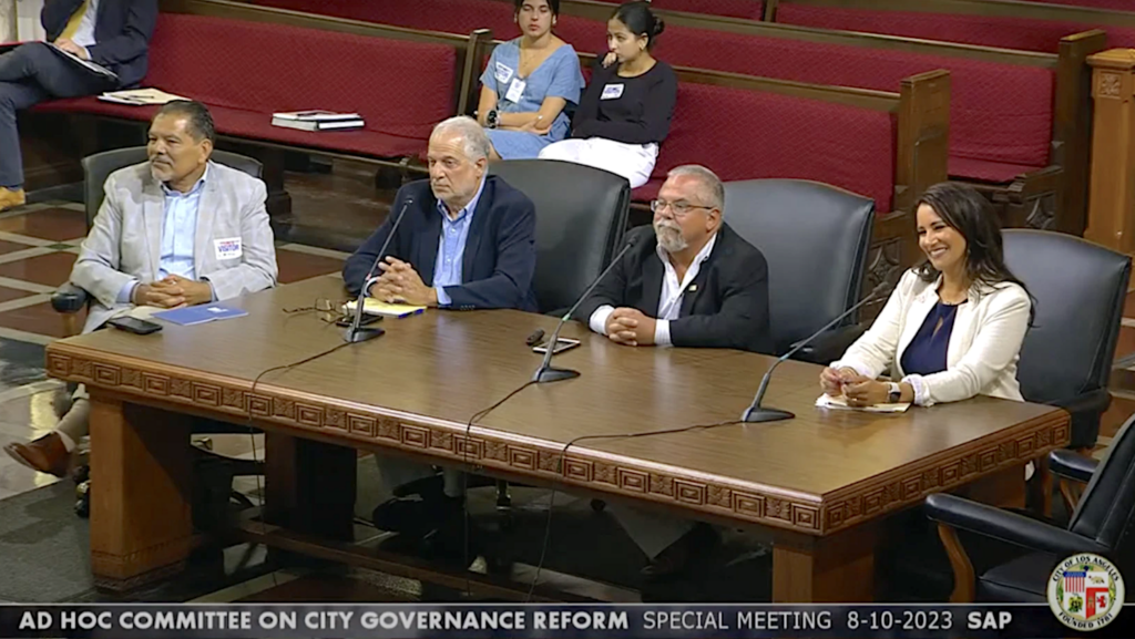 three men and one women at large wooden desk with microphones