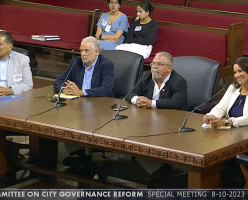 three men and one women at large wooden desk with microphones