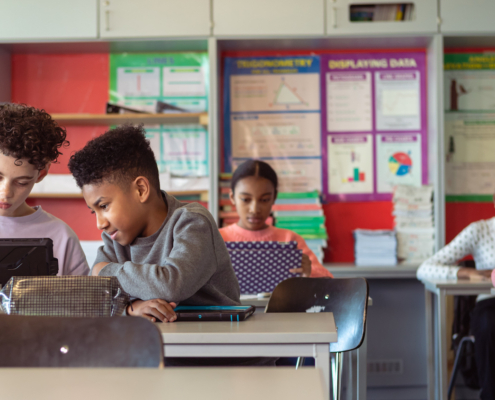 Multiracial group of adolescent kids sit at desks in their classroom and use tablet computers. Selective focus on two boys who are working on an assignment together.