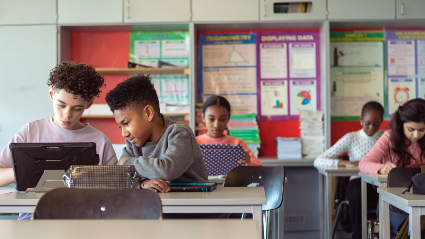 Multiracial group of adolescent kids sit at desks in their classroom and use tablet computers. Selective focus on two boys who are working on an assignment together.