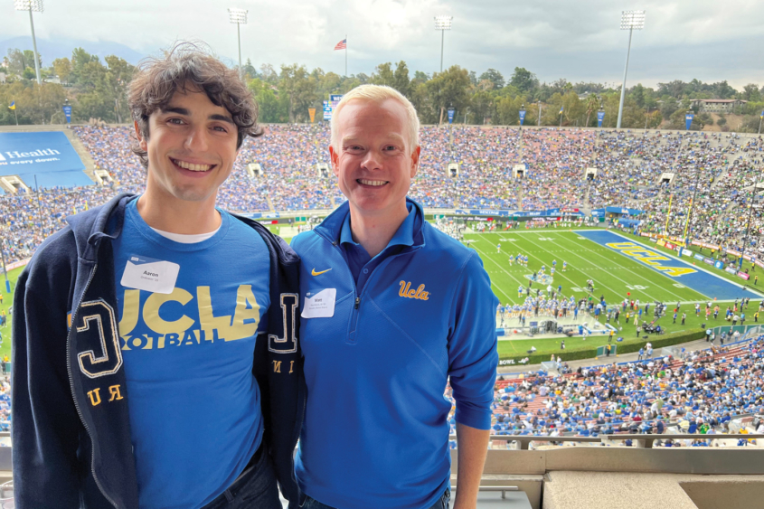 two young men smile in stands as UCLA football game goes on behind them