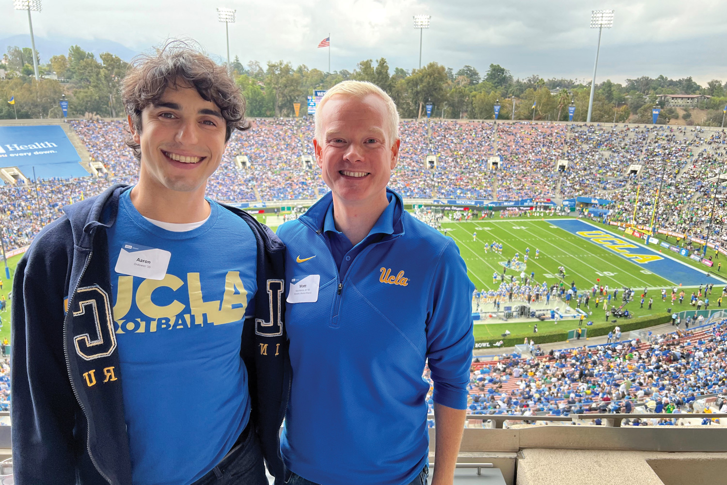 two young men smile in stands as UCLA football game goes on behind them
