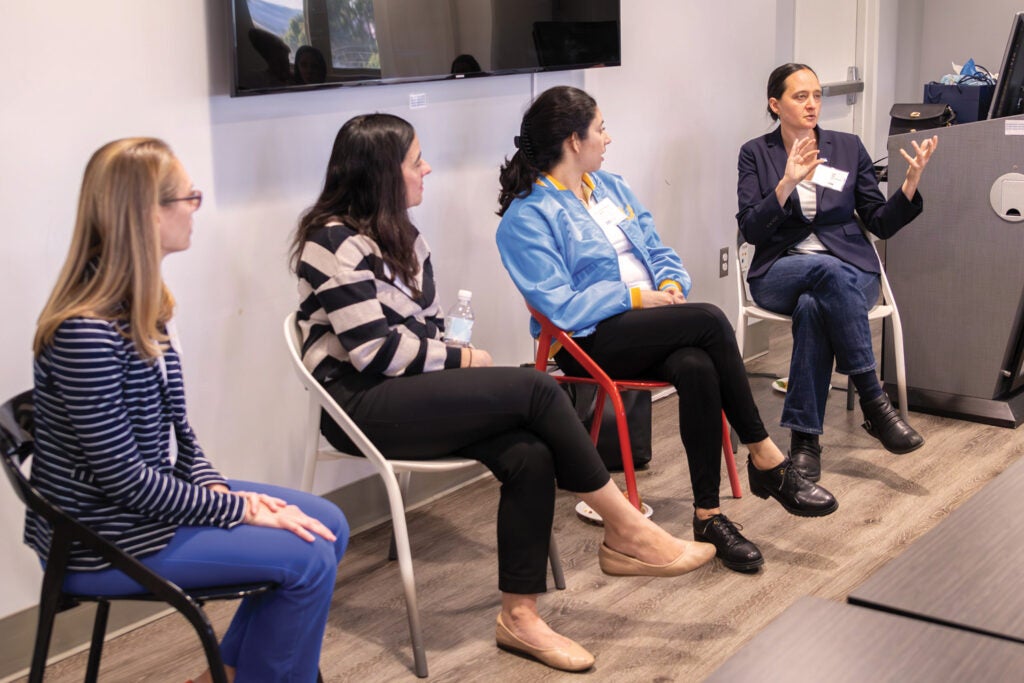 four women are seated in chairs, with three listening as the fourth on far right gestures while talking|portrait photo of Amanda Morrall|portrait photo of Leslea Meyerhoff