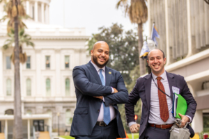 two men in suits pose with state Capitol building behind them