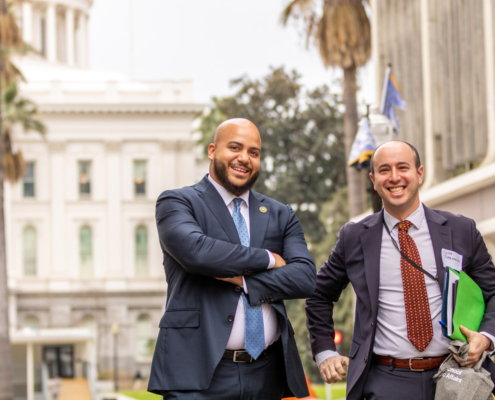 two men in suits pose with state Capitol building behind them