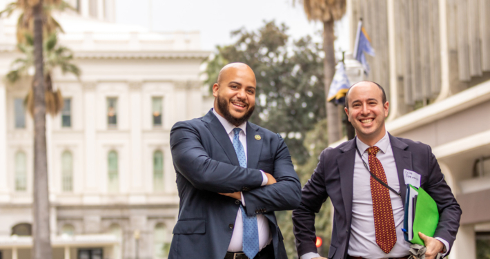 two men in suits pose with state Capitol building behind them