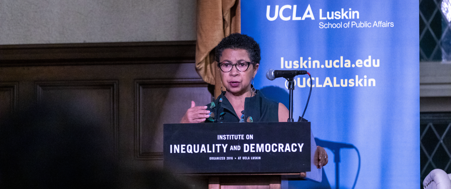 Black woman wearing glasses speaks from a podium with a UCLA Luskin backdrop behind her
