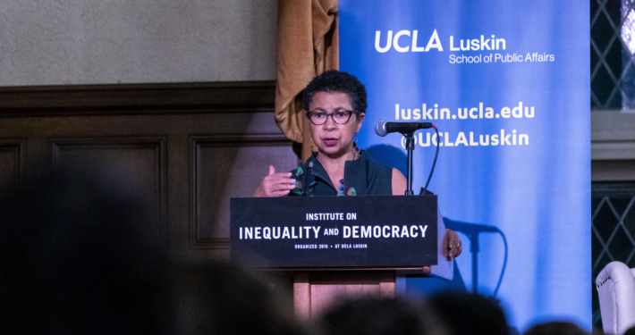 Black woman wearing glasses speaks from a podium with a UCLA Luskin backdrop behind her