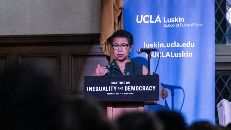 Black woman wearing glasses speaks from a podium with a UCLA Luskin backdrop behind her