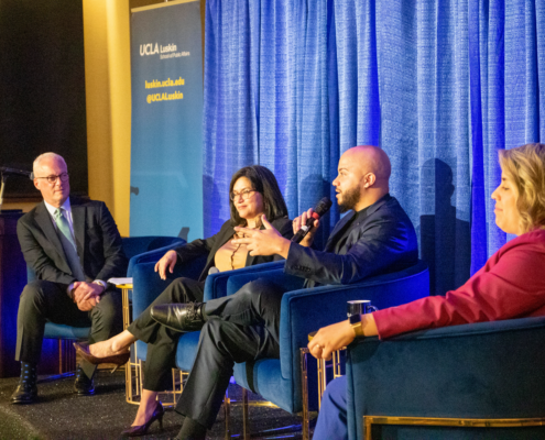 four people in business attire site on a stage and chat with a blue background behind them