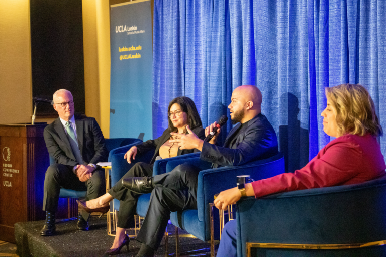 four people in business attire site on a stage and chat with a blue background behind them|||||