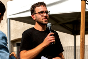 young man wearing glasses speaks into a microphone he holds in his right hand