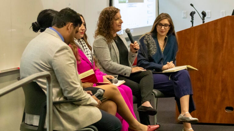 five people in business attire sit on a stage as a woman with curly brown hair speaks into a microphone