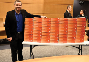 man in dark suit rests one arm on a huge stack of books on a folding table