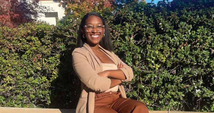 Black woman dressed in beige sweater and brown pants folds arms across chest