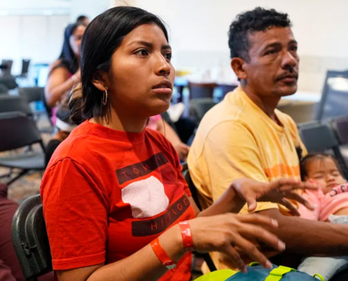 two adults and an infant sit in chairs at an orientation for new immigrants